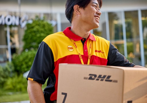 DHL Express courier in a red and yellow uniform holding a large brown DHL Express Box 7 outdoors near a building.
