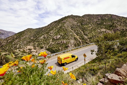 Yellow DHL delivery van driving along a winding mountain road with vibrant orange flowers in the foreground and hills in the background.